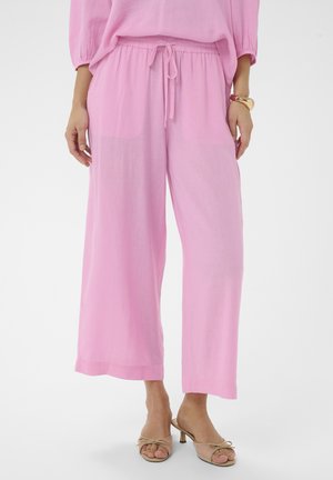Kobieta w luźnych, jasnoróżowych spodniach z troczkami, beżowych sandałach na niskim obcasie z odkrytymi palcami oraz dopasowanym różowym topie, stoi na białym tle.