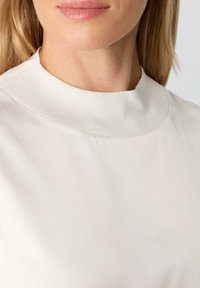Jasno beżowa bluzka z wysokim, prążkowanym dekoltem, gładką fakturą i prostym, minimalistycznym designem, odpowiednia na różne okazje.