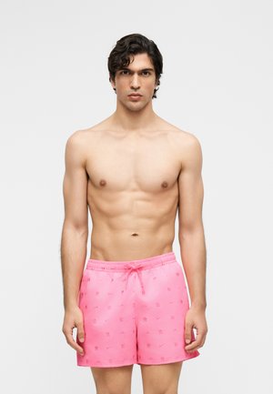 Giovane uomo con capelli ricci scuri che indossa pantaloncini da bagno rosa acceso con fantasia, in piedi davanti a uno sfondo chiaro e uniforme.