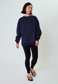 Felpa navy a maniche lunghe, con polsini e orlo ribattuti, abbinata a leggings neri e infradito neri, su uno sfondo chiaro.