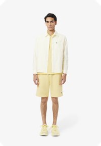 Camicia button-up a righe giallo chiaro sopra una maglietta coordinata e pantaloni corti. L'outfit presenta una texture morbida e piccoli dettagli con logo.