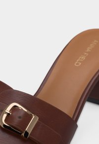Mule à talon en cuir marron avec une boucle en métal doré sur la sangle et une semelle intérieure beige présentant un logo imprimé discret.