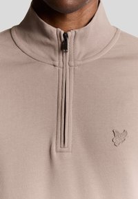 Beige sweatshirt met ritssluiting, gemaakt van een gestructureerde stof. Kenmerkt zich door een zwarte rits en een klein geborduurd logo aan de onderkant rechts.