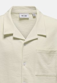 Camicia a maniche corte color crema con tessuto testurizzato, colletto e taschino sul petto; presenta tre bottoni bianchi sul davanti.