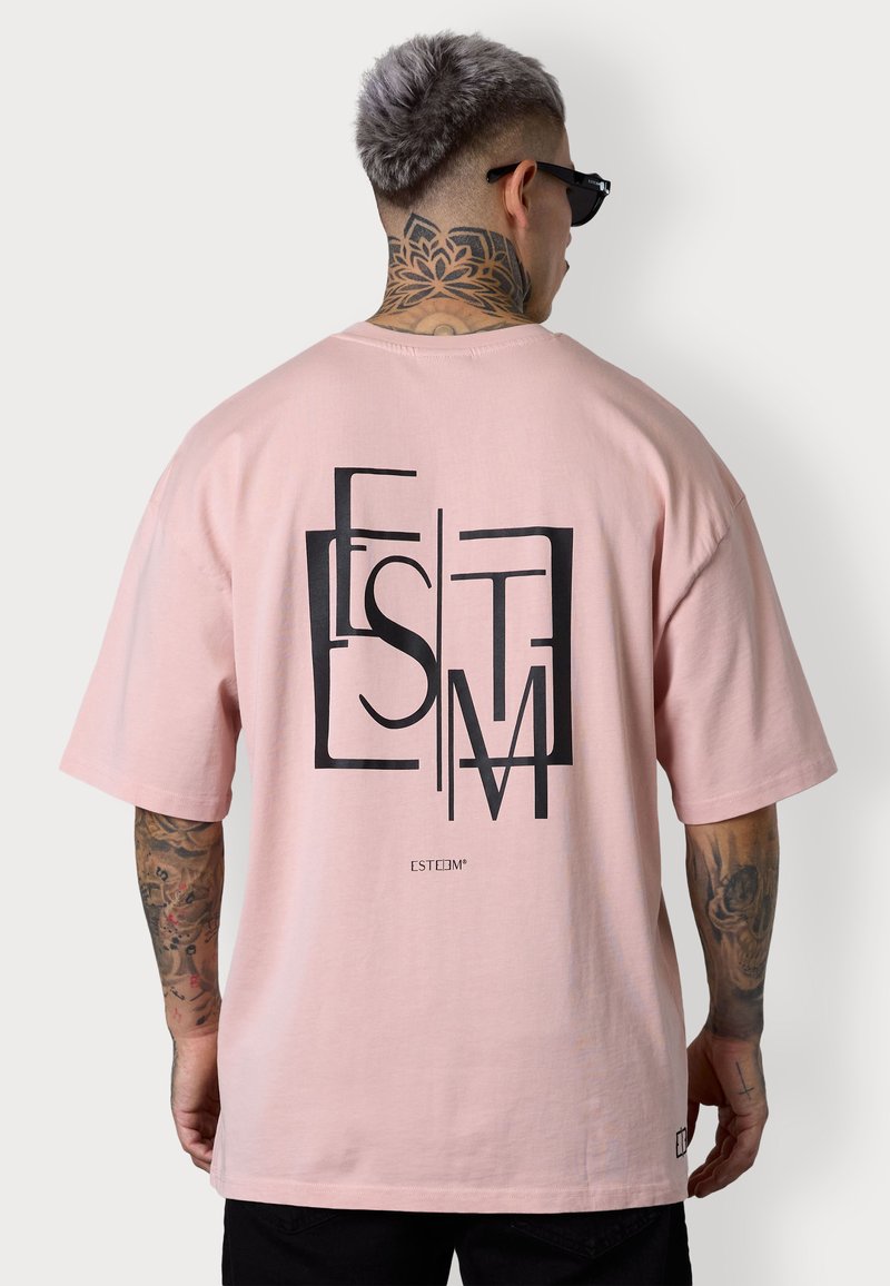 T-shirt oversized rose clair en coton ; présente un grand design graphique noir au dos et des manches courtes et amples.