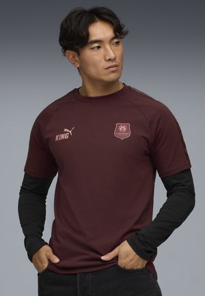 Bordeaux kortærmet skjorte med rund hals, som har sorte langærmede bluser nedenunder og et logo med et løveemblem på brystet.