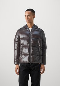 Pufferjack in glanzend donkergrijs materiaal, gewatteerd ontwerp, frontale rits, twee zijzakken en een capuchon. Gedragen over een wit shirt.