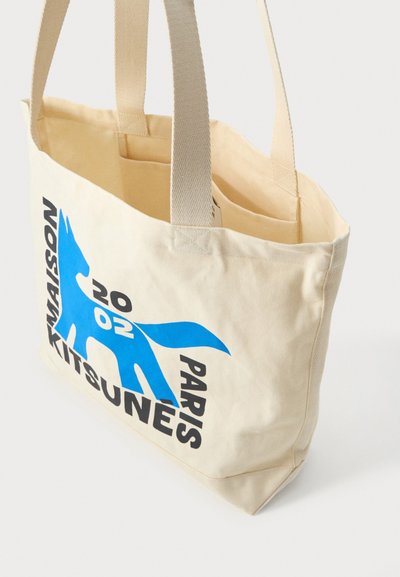 Geantă bej cu două mânere, cu un grafic cu o vulpe albastră și text negru pe lateral cu inscripția „MAISON KITSUNÉ PARIS 2002”.