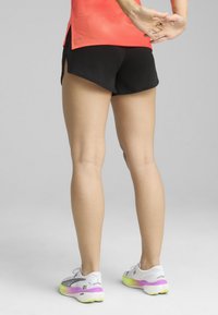 Pantalones cortos deportivos negros combinados con una camiseta de color naranja brillante, con un corte relajado y aberturas laterales, llevados con zapatillas de correr blancas y en degradado pastel.