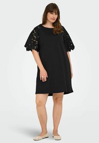 Woman wearing a black knee-length dress with lace short sleeves and beige flat shoes, standing against a plain light background.