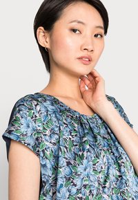 Blusa de mangas curtas com padrão floral apresentando flores azuis e verdes sobre um fundo escuro, com um decote franzido e uma textura de tecido suave.