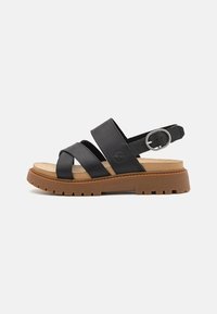 Mustat nahka sandaalit, joissa on leveät ristikkäiset hihnat, takana solki ja vaalea ruskea pehmeä pohja, jossa on teksturoitu ruskea pohja.