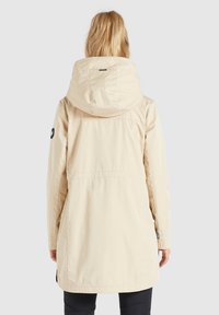 Jasna beżowa wodoodporna kurtka z kapturem, długimi rękawami i ściągaczem w talii, z czarną naszywką z logo na lewym rękawie.