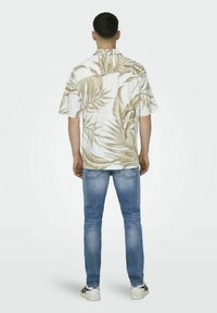 Cămașă cu mânecă scurtă, albă, cu un model de frunze de palmier bej, asortată cu blugi deschiși la culoare și adidași albi, văzută din spate.