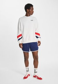 Man in een wit sweatshirt met rode en marineblauwe strepen, marineblauwe shorts, witte sokken met zwarte chevrons, en rode sneakers, staande en naar links kijkend.