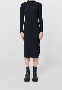 Robe noire en maille côtelée à manches longues, col rond, buste ajusté et ourlet mi-long. Portée avec des bottines noires à semelles épaisses.