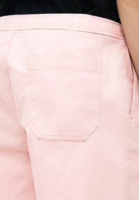 Ροζ παντελόνι με τετράγωνη πίσω τσέπη και εμφανή ραφή, φορεμένο με μαύρο τοπ, εμφανίζοντας πλάγια και πίσω όψη της περιοχής της μέσης.