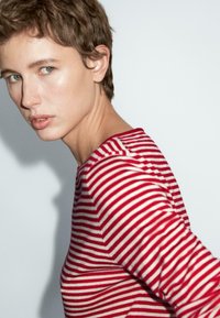 Persoon met kort haar, gekleed in een rood-wit gestreept shirt met lange mouwen, draait het hoofd iets naar de camera tegen een effen achtergrond.