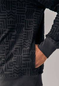 Svart texturerad sweatshirt med upphöjda geometriska mönster, ribbade muddar och en sidoficka. Mjuk, plyschliknande material med en modern design.