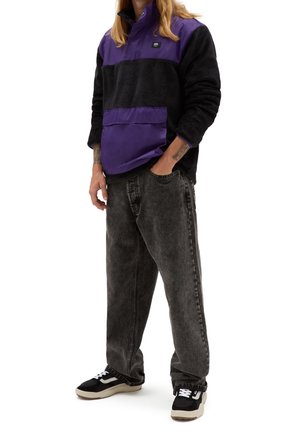 Person wearing black and purple fleece pullover, loose faded black jeans, and black and white sneakers, standing with one hand in pocket.