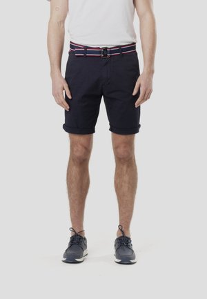 Tengerészkék pamut short felhajtott szegéllyel, sokszínű csíkos övvel. A modell egyszínű fehér pólót és tengerészkék tornacipőt visel.