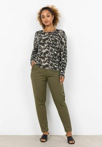 Langärmliges Oberteil in Schwarz und Creme mit Muster, kombiniert mit olivgrünen Hosen und schwarzen offenen Schuhen. Weiche Textur, lockere Passform und sichtbare Taschen.