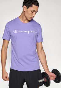 Μωβ κοντομάνικη μπλούζα με κεντημένο λευκό λογότυπο "Champion". Φοριέται από μοντέλο που κρατάει μια μαύρη αλτήρα, συνδυασμένη με μαύρο σορτς.