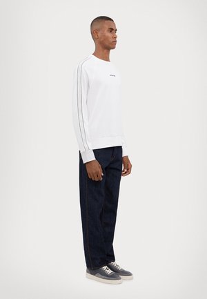 White long-sleeve sweatshirt with black side stripes and logo, paired with dark denim trousers and gray sneakers with white soles.