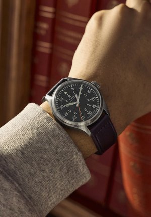 Käekell musta sihverplaadi ja valgete märkidega inimese randmel, mis näitab aega 11:15, taustal udused punased raamatud.