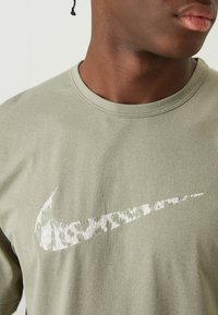 Olajzöld Nike póló, nagy, textúrált swoosh logóval fehér színben, finom mintázattal; rövid ujjú és kerek nyakú.