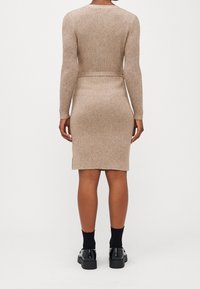 Robe en maille beige côtelée avec une silhouette ajustée et des manches longues. La taille est cintrée par une ceinture assortie. Des chaussures noires et des chaussettes complètent le look.