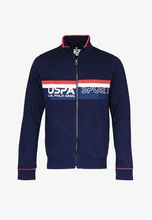 Tummansininen vetoketjullinen takki, joka on valmistettu pehmeästä kankaasta ja jossa on korkea kaulus. Takissa on punaisia ja valkoisia vaakasuoria raitoja sekä U.S. Polo Assn. -logo edessä.