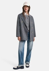 Grijze dubbelgeknoopte wollen blazer met gouden knopen, gedragen over een grijs overhemd met knopen. Lichte blauwe casual jeans en marineblauwe instappers met witte accenten.