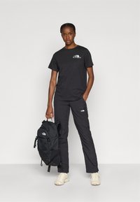 Μαύρο μπλουζάκι με ένα μικρό λογότυπο, μαύρο παντελόνι και μαύρο σακίδιο πλάτης. Το μοντέλο φορά ανοιχτόχρωμα αθλητικά παπούτσια και στέκεται μπροστά σε ένα απλό φόντο.