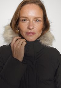 Juodas pūkuotasis švarkas su dirbtiniu kailiniu ant gaubto. Medžiaga atrodo blizgi su siuvinėtomis dalimis, suteikianti šiltą ir izoliuotą dizainą.