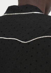 Czarna koszula z materiału, ozdobiona wypukłymi, teksturowanymi kropkami, z wygiętą linią srebrnych rhinestones wzdłuż dekoltu, co dodaje dekoracyjnego akcentu.