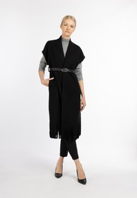 Zwarte franjesvest met een riem met een ronde gesp, gelaagd over een grijze polo en gecombineerd met zwarte strakke broeken en hoge hakken.