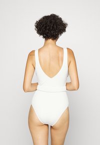 Maillot de bain une pièce blanc avec un tissu texturé, des bretelles larges, un dos en V profond et une taille ceinturée.