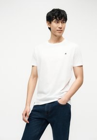 Ung mann iført en hvit kortermet T-skjorte med en liten logo på brystet og blå jeans, poserer med en hånd i lomma.