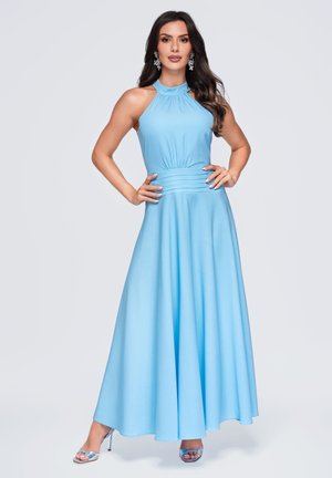 Femme portant une robe bleu clair sans manches à col halter et des sandales argentées à talons hauts, debout les mains sur les hanches.