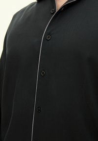 Camicetta nera a maniche corte realizzata in tessuto liscio con colletto classico, dotata di bottoni neri e piping grigio chiaro a contrasto lungo i bordi.