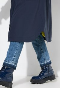 Manteau imperméable marine avec doublure jaune, associé à un jean bleu déchiré, et des bottes militaires navy brillantes avec de épaisses semelles noires.