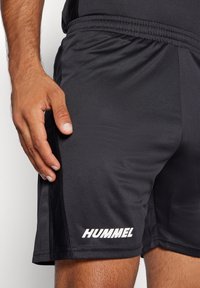 Homme portant un short de sport noir avec le logo « HUMMEL » sur la jambe gauche, debout avec la main gauche détendue à côté du short.