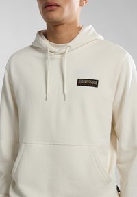 Sudadera beige hecha de tela suave, presenta un bolsillo canguro frontal y un parche negro con el logo de la marca en el pecho. Incluye capucha con cordón.