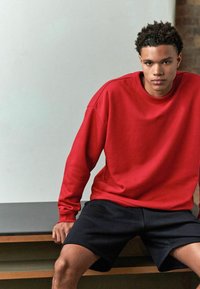 Rød sweatshirt med ribbet udskæring og lange ærmer, parret med sorte shorts. Glat stof med en afslappet pasform og minimal detaljer.