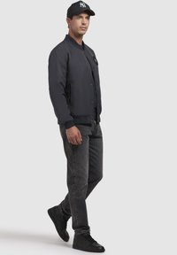 Jachetă de bombardier bleumarin cu o aplică logo, blugi negri și adidași negri cu glezna înaltă. Textură netedă; design ajustat cu manșete și tiv cu nervuri.