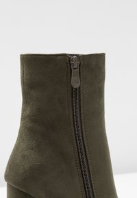 Botte de cheville en daim vert olive avec un haut col et un détail de fermeture éclair sur le côté, mettant en valeur une texture douce et une forme effilée.
