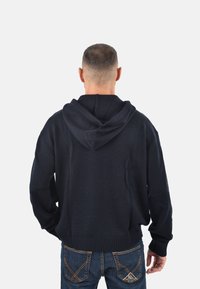 Felpa con cappuccio in maglia blu navy con finitura testurizzata, dotata di cappuccio con coulisse e polsini a costine. Indossata con jeans blu, vista posteriore.