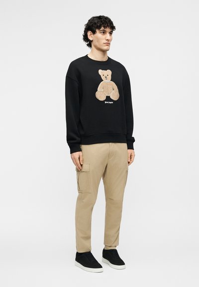 Άνδρας που φοράει μαύρο φούτερ με γραφικό αρκουδάκι και μπεζ παντελόνι cargo, στέκεται μπροστά σε απλό ανοιχτό φόντο.