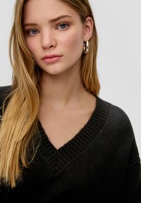 Schwarzer V-Ausschnitt-Pullover mit geripptem Kragen; das Model hat lange, wellige blonde Haare und trägt silberne Creolen; neutraler Hintergrund.